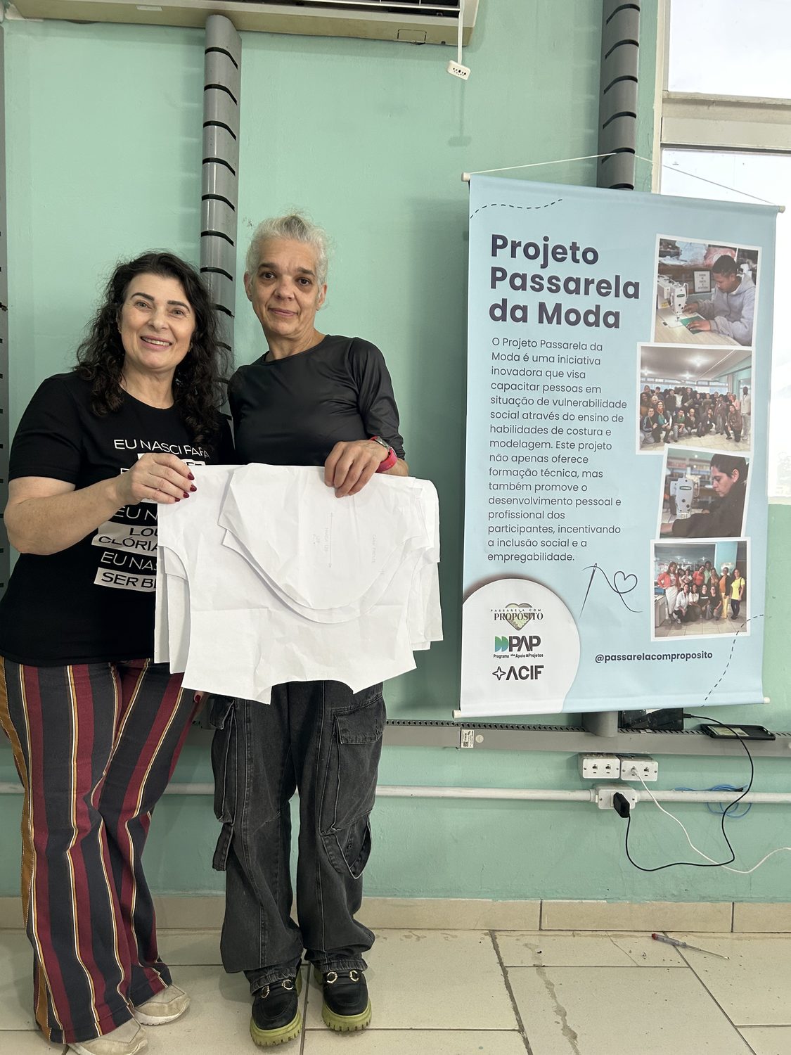 Duas participantes sorrindo, segurando uma peça branca que confeccionaram, ao lado do banner do Projeto Passarela da Moda
